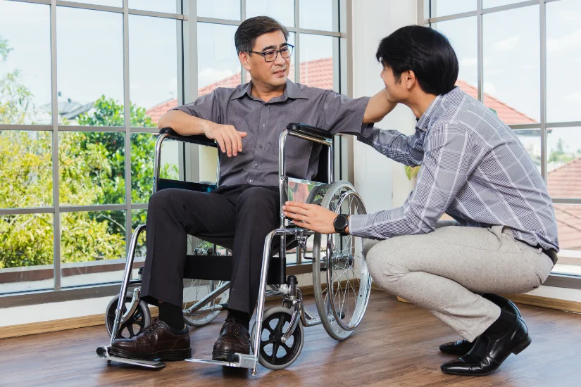 Asian senior disabled businessman in wheelchair discuss interacting together with the team in the office. The old man in a wheelchair and his young son talking to and comforting bound father