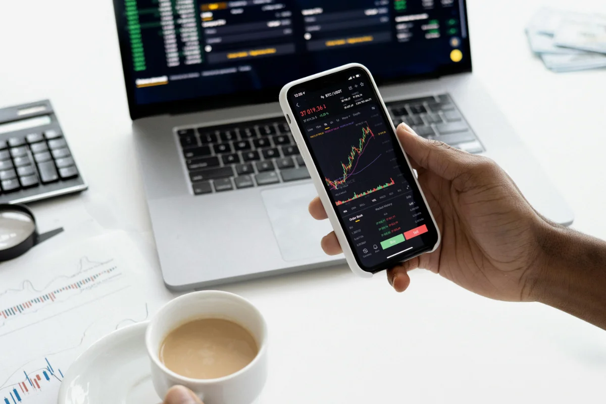 Hand holding mobile phone with investment charts, and laptop, cup, calculator and paper at the background.