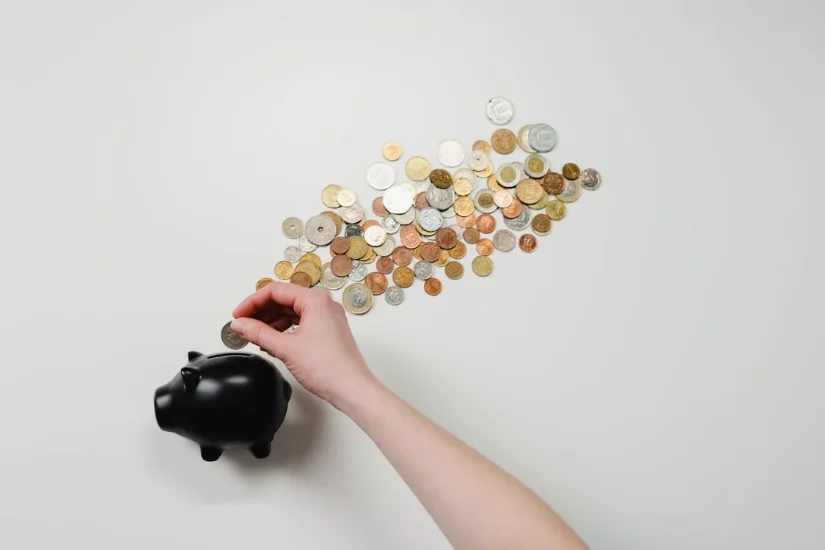 Hand holding coin attempt to insert into piggy bank, with lots of coins at background.