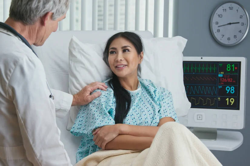 Supportive and caring older doctor with hand on shoulder of recovering female patient in hospital bed. Japanese woman getting good news from senior Caucasian physician