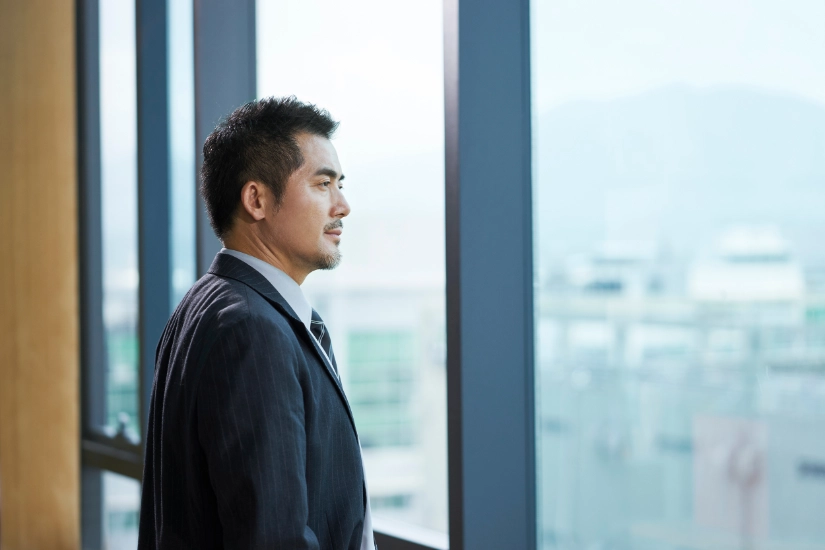 portrait of a mature asian business man standing in front of window looking out and thinking